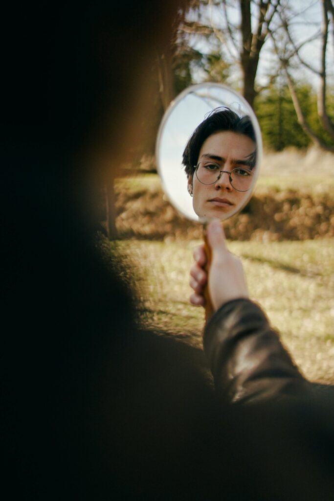 young man standing in woods holding a mirror. The reflection of his face is in the mirror.