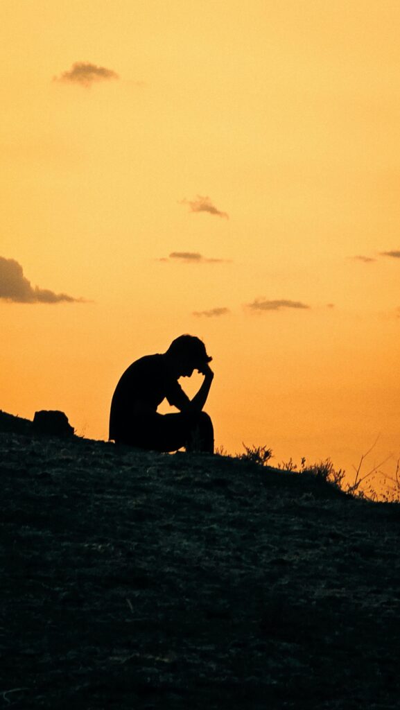 Silhouette of a person sitting alone at sunset with their head in their hand, representing emotional exhaustion, depression, anxiety, or deep reflection.